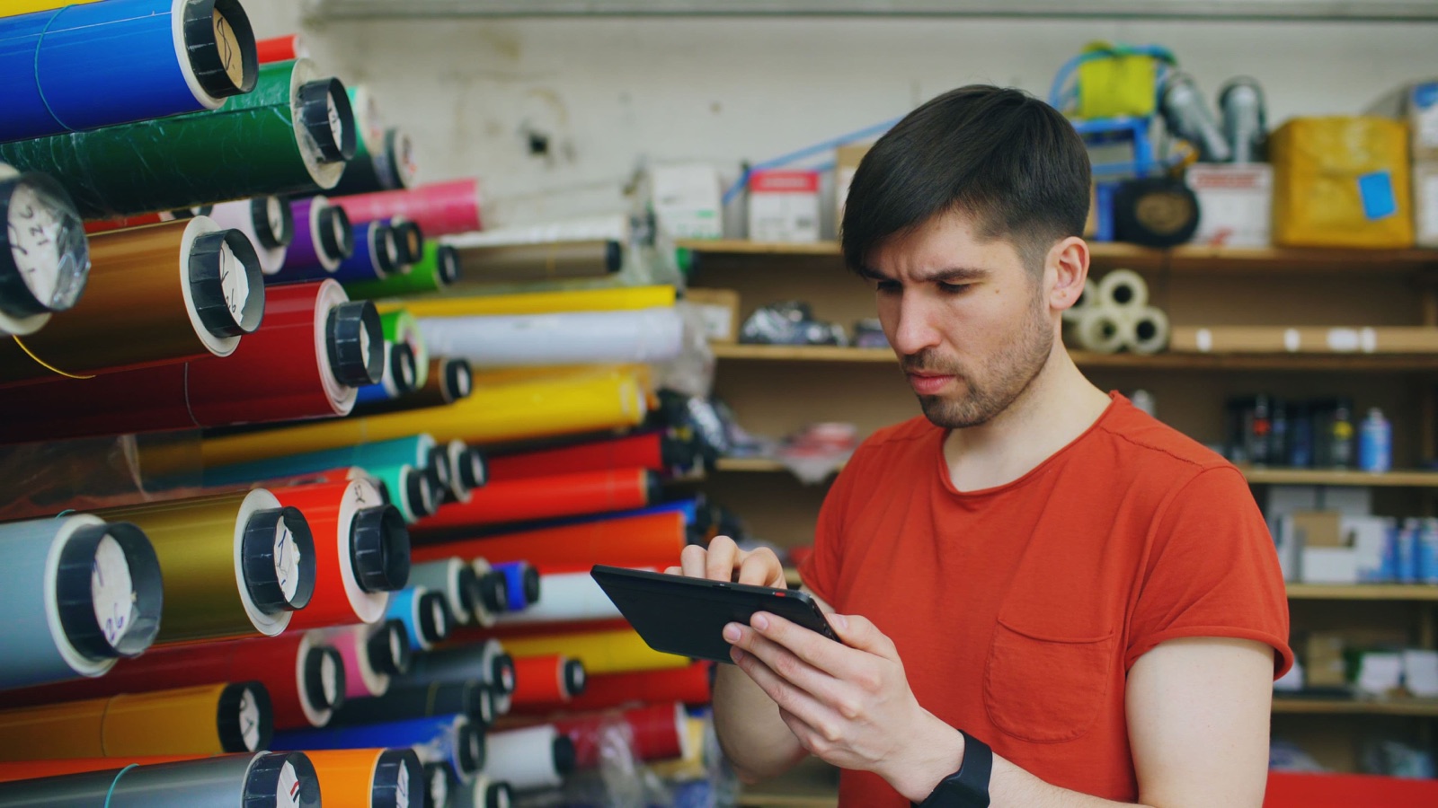 Small-business owner in a red t-shirt checking inventory on a tablet inside his materials shop, surrounded by colourful vinyl rolls — the kind of European small-business operator looking for ways to cut costs without cutting people.